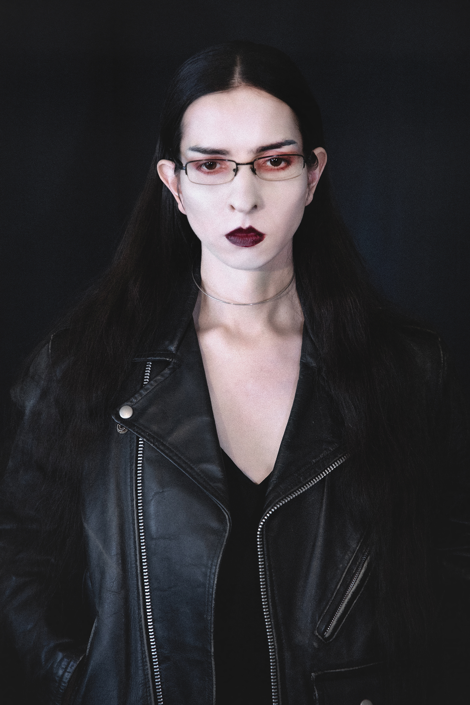 Portrait of a female model with pale skin and rust colored eye and lip makeup in a leather jacket looking downwards against a dark backdrop.